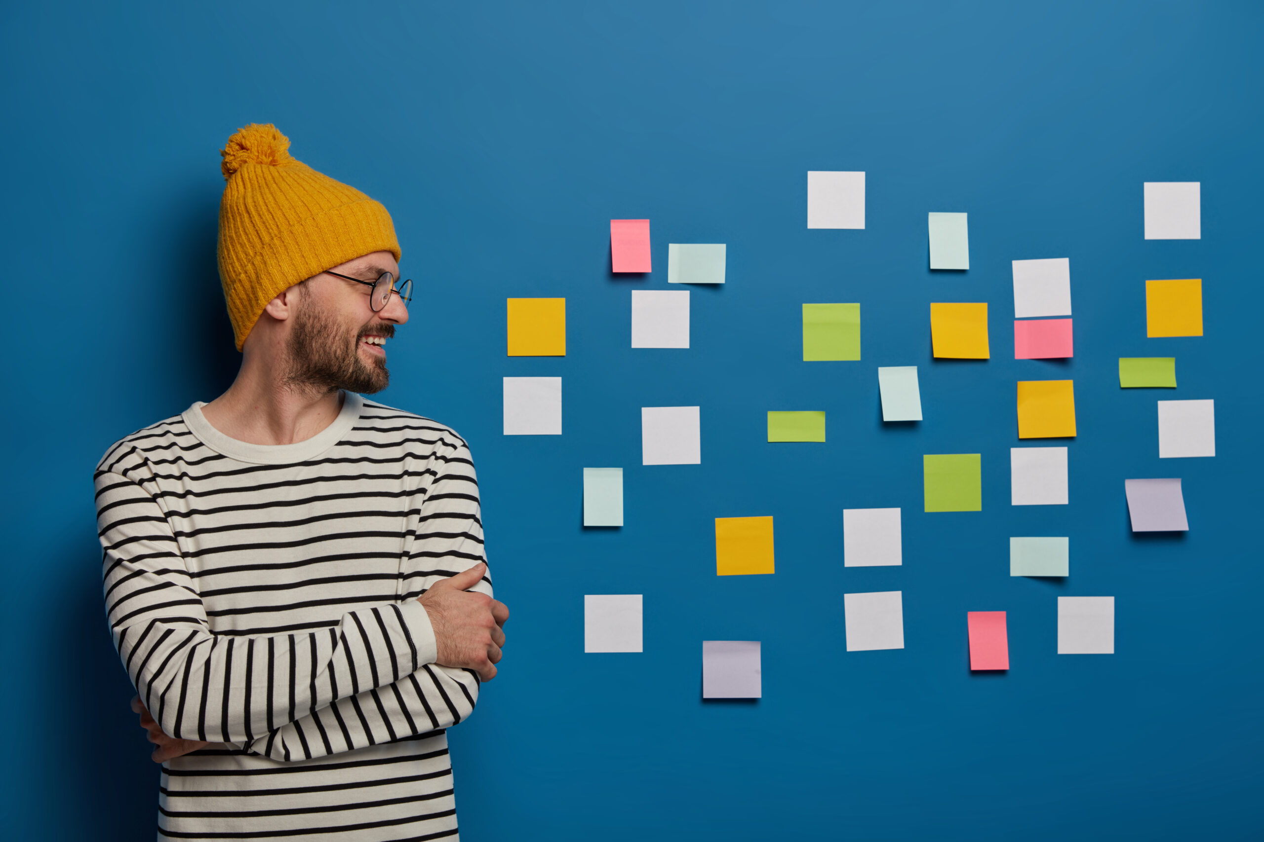 Happy creative male freelance worker stands with hands crossed, wears yellow hat and striped jumper, looks on right side stands at working place, sticky notes for leaving reminder
