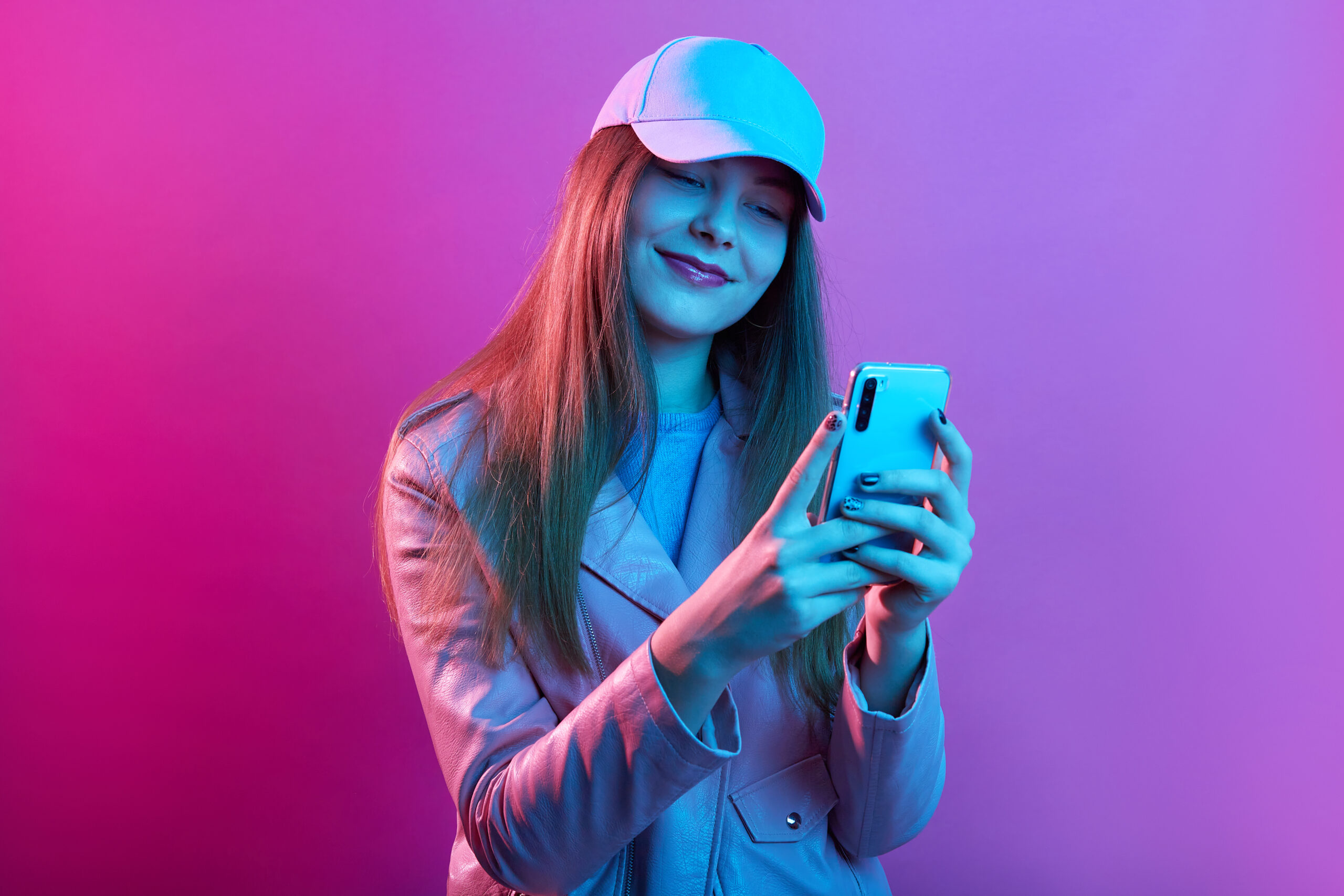 Portrait of young beautiful fashionable model wearing leather baker and baseball cap, holding smart phone in hands, texting with friends or checking social networks